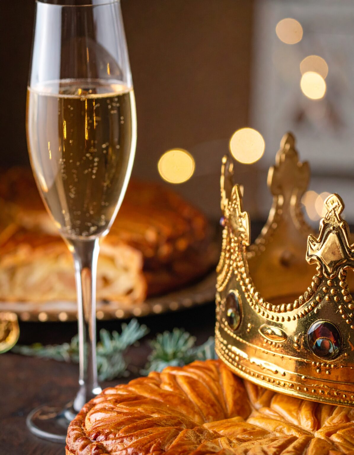 Flûte de champagne remplie de bulles dorées posée à côté d’une galette des rois croustillante, surmontée d’une couronne dorée ornée de pierres, avec des lumières floues en arrière-plan créant une ambiance chaleureuse et festive.
